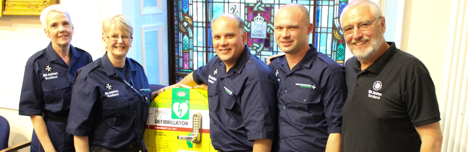 A group of people in St John Scotland uniform pose with a defibrillator cabinet