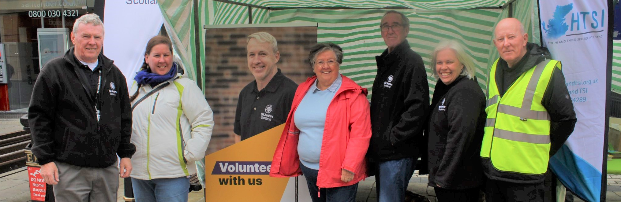 A group of six volunteers pose in front of a pop up banner and stand