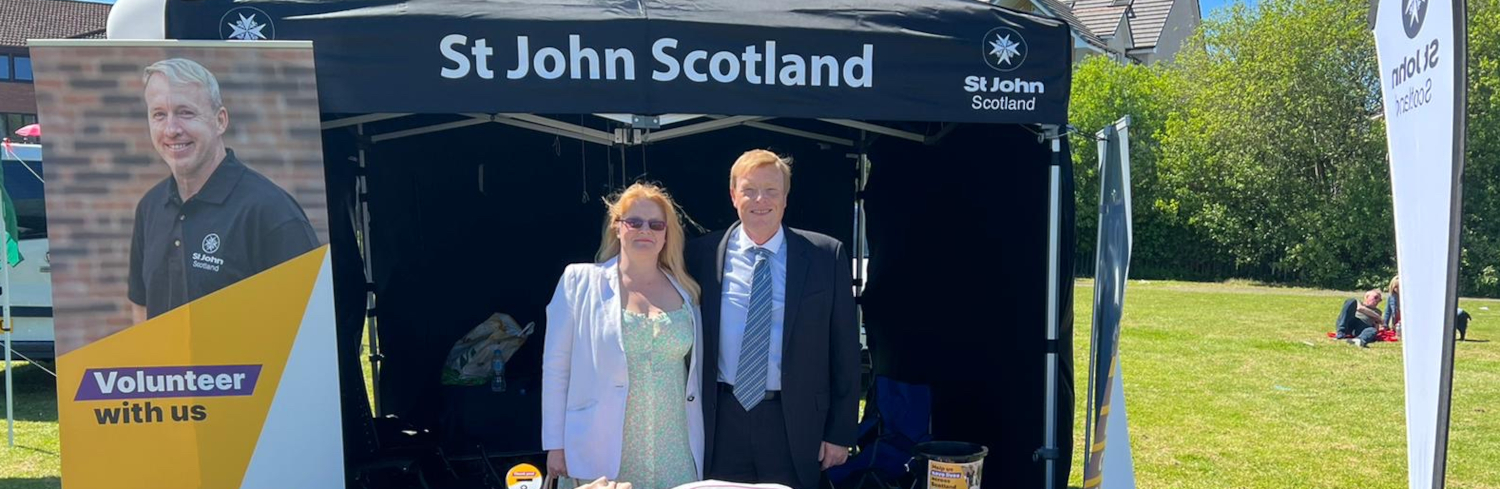 Two St John Scotland volunteers stand in front of a gazebo next to a sign that says Volunteer With Us