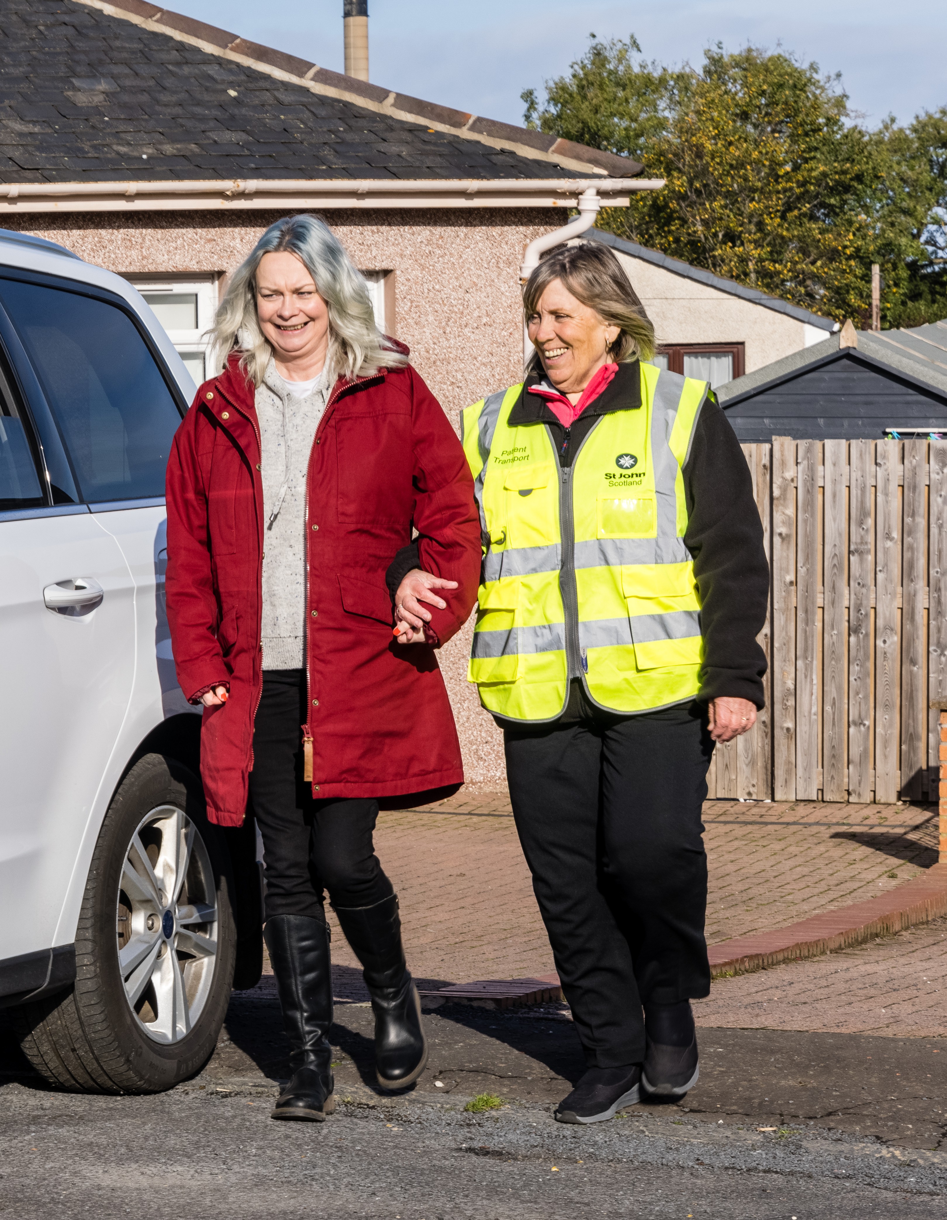 2 women walk arm in arm next to a parked car. one woman is in a high visibility jacket
