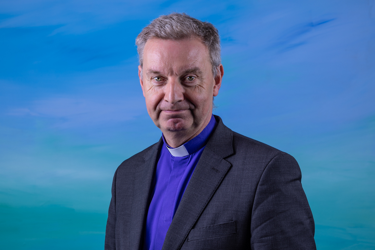 A man in a suit and clerical collar in front a blue background