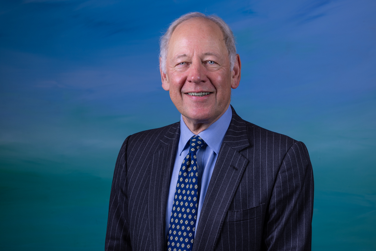 A man in front of a blue background. He is wearing a striped suit