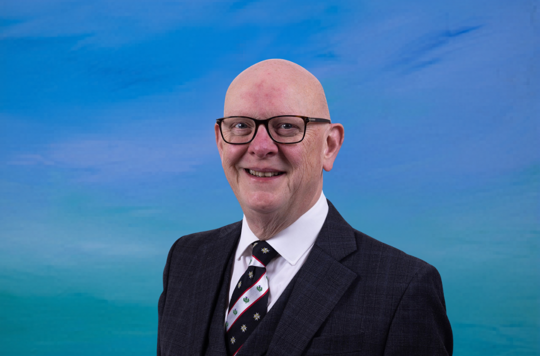 A bald man with glasses sits in front of a blue background. He has a suit and a St John Scotland tie on