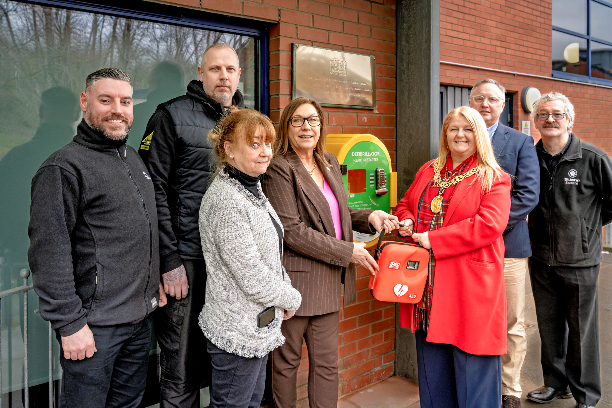 7 people (4 men and 3 women) stand around a defibrillator case. The defibrillator is being handed to one of the women