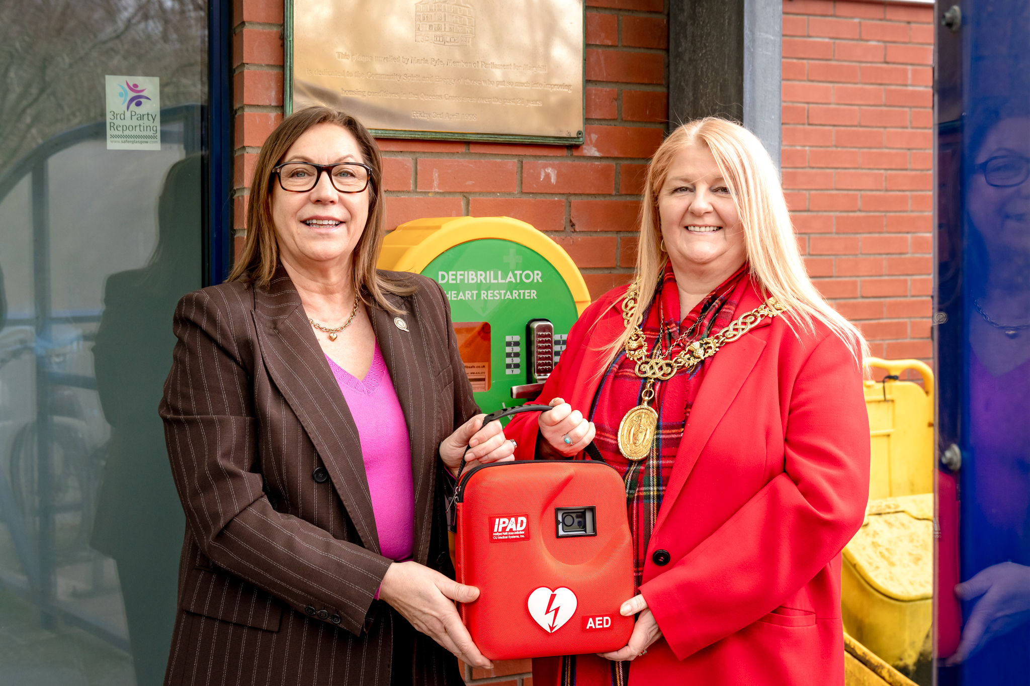 2 women stand in front of a defibrillator case. They are holding the defibrillator in between them