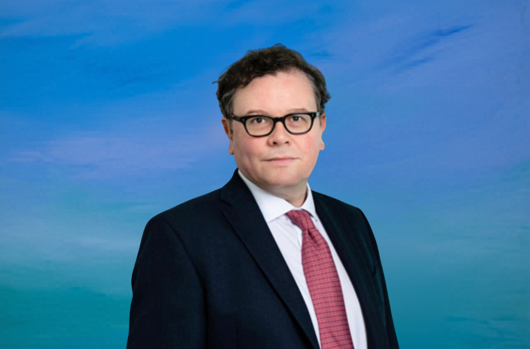 A man with glasses in front of a blue background. He is wearing a suit.