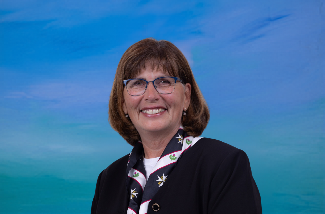 A woman with shoulder length brown hair and glasses smiles at the camera in front of a blue background