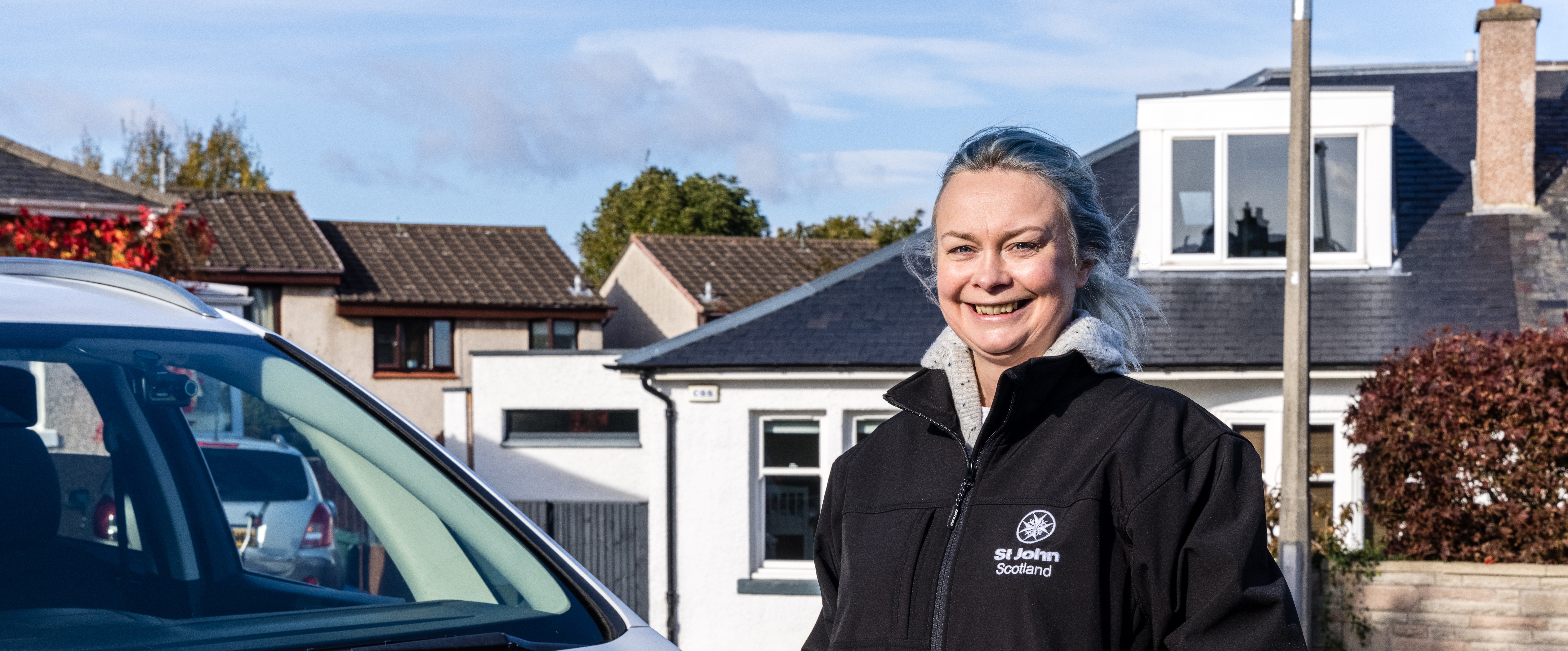 A woman stands in front of a white car with the St John Scotland logo on it. She is wearing a St John Scotland fleece