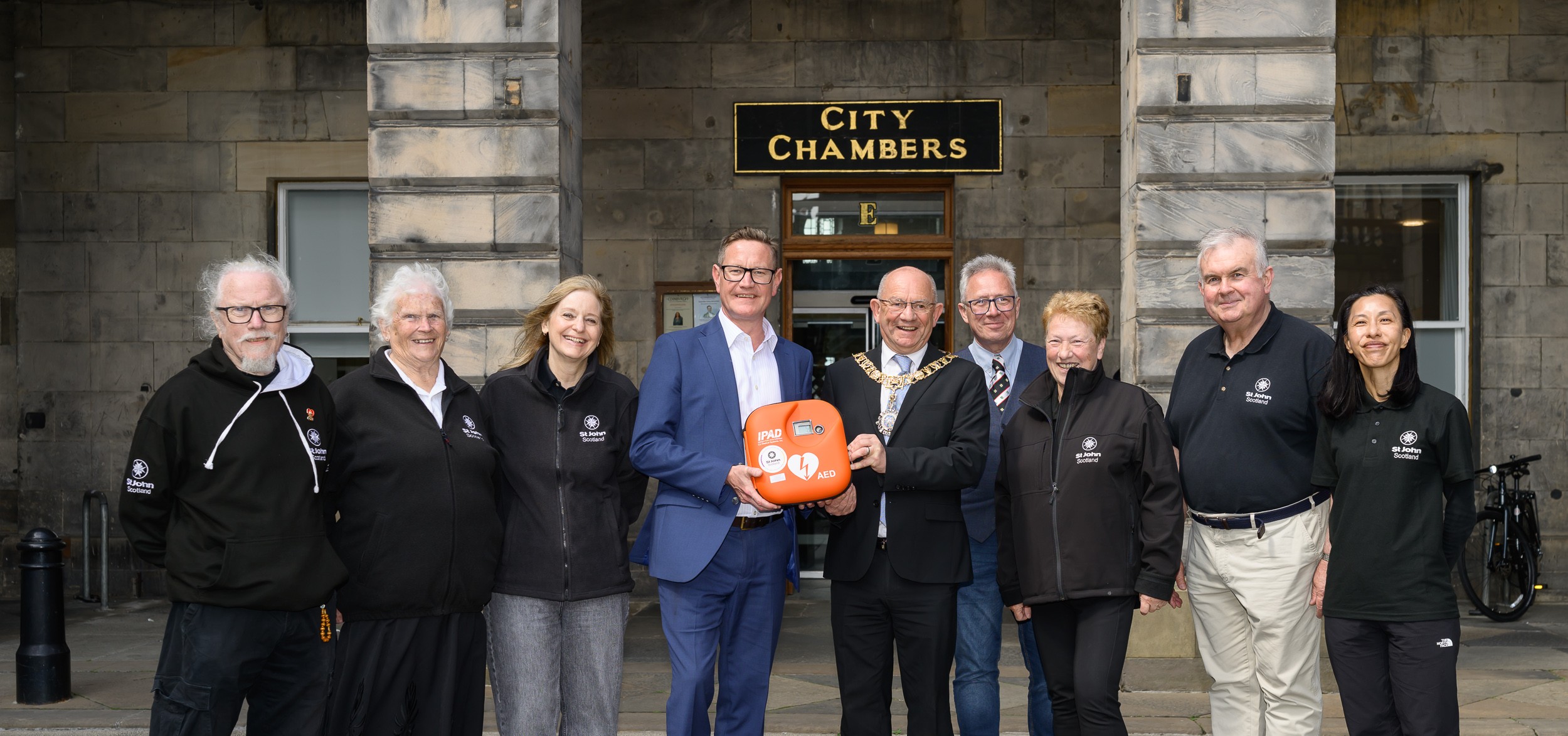 A group of 9 people stand in a line smiling. In the middle 2 men are holding a Public Access Defibrillator