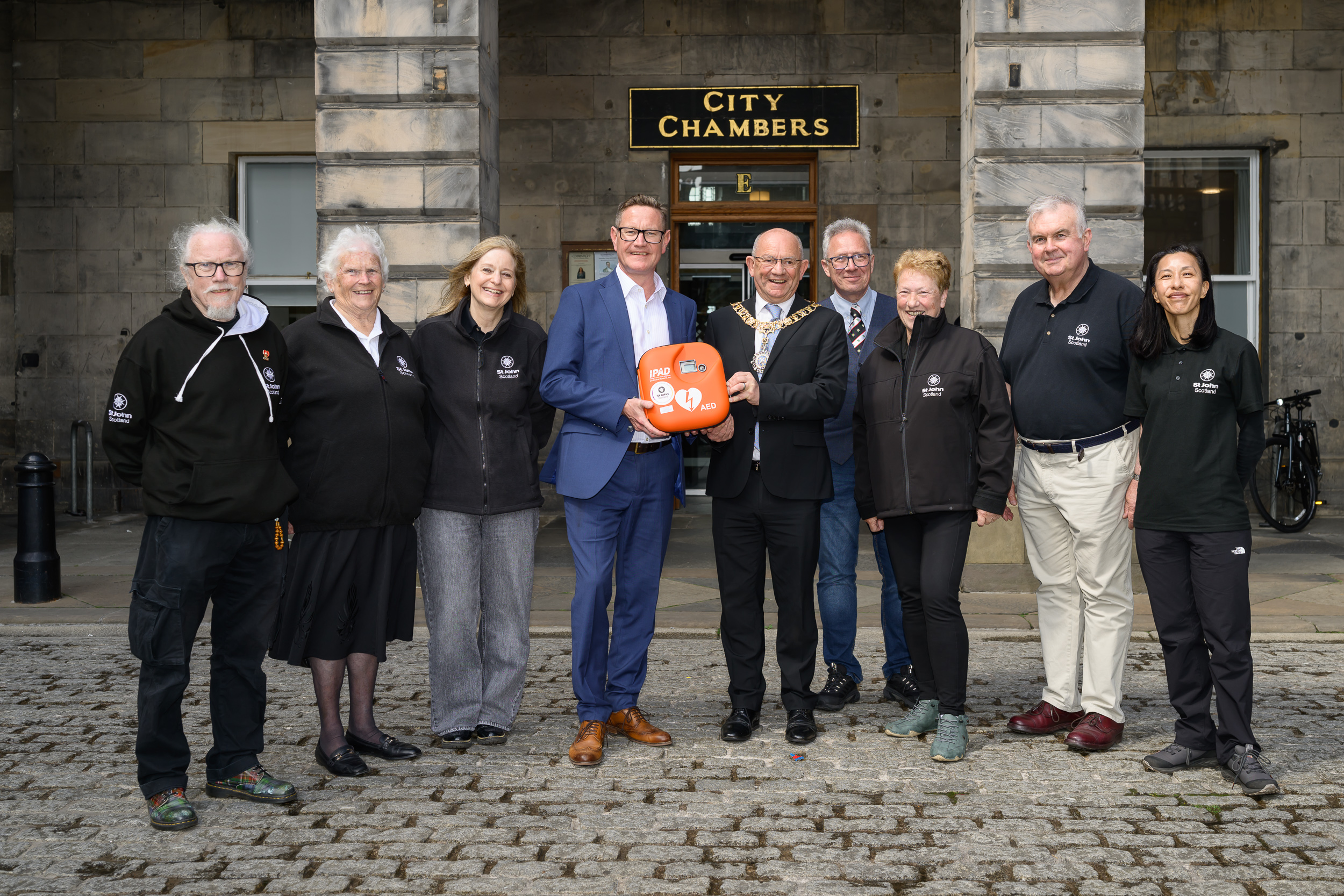 A group of 9 people stand in a line smiling. In the middle 2 men are holding a Public Access Defibrillator