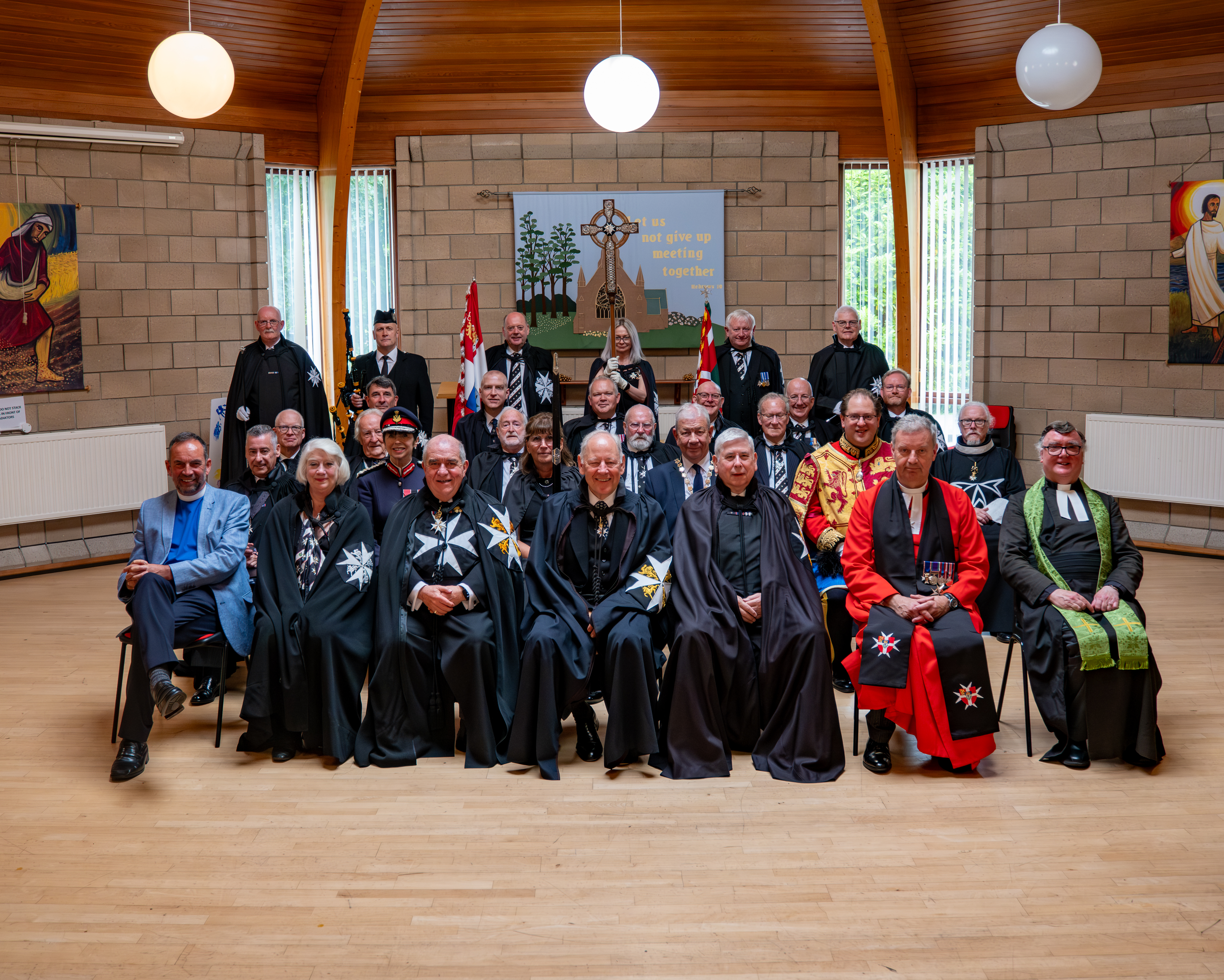 A large group of people sit and stand in front of the camera in rows. Many of them are dressed in traditional St John robes