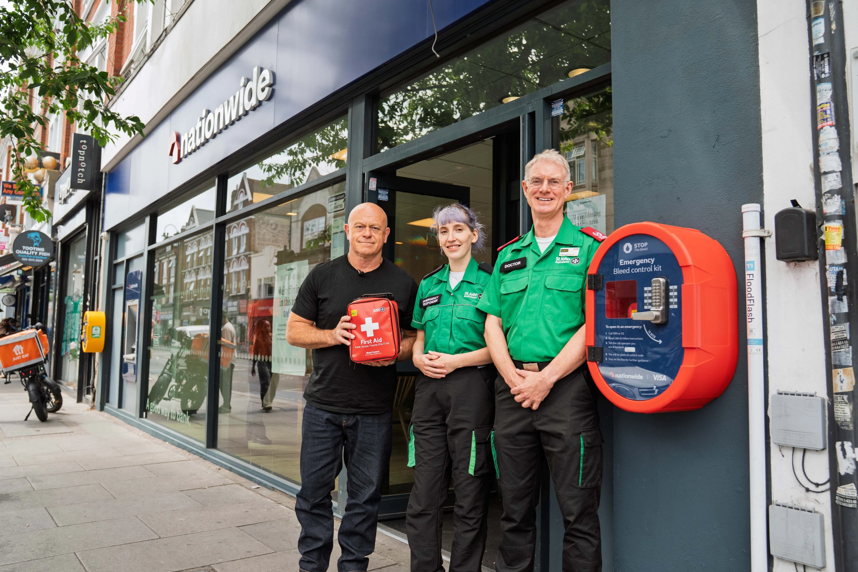 Ross Kemp stands with 2 people in St John Ambulance uniforms outside a Nationwide branch. They are holding a defibrillator