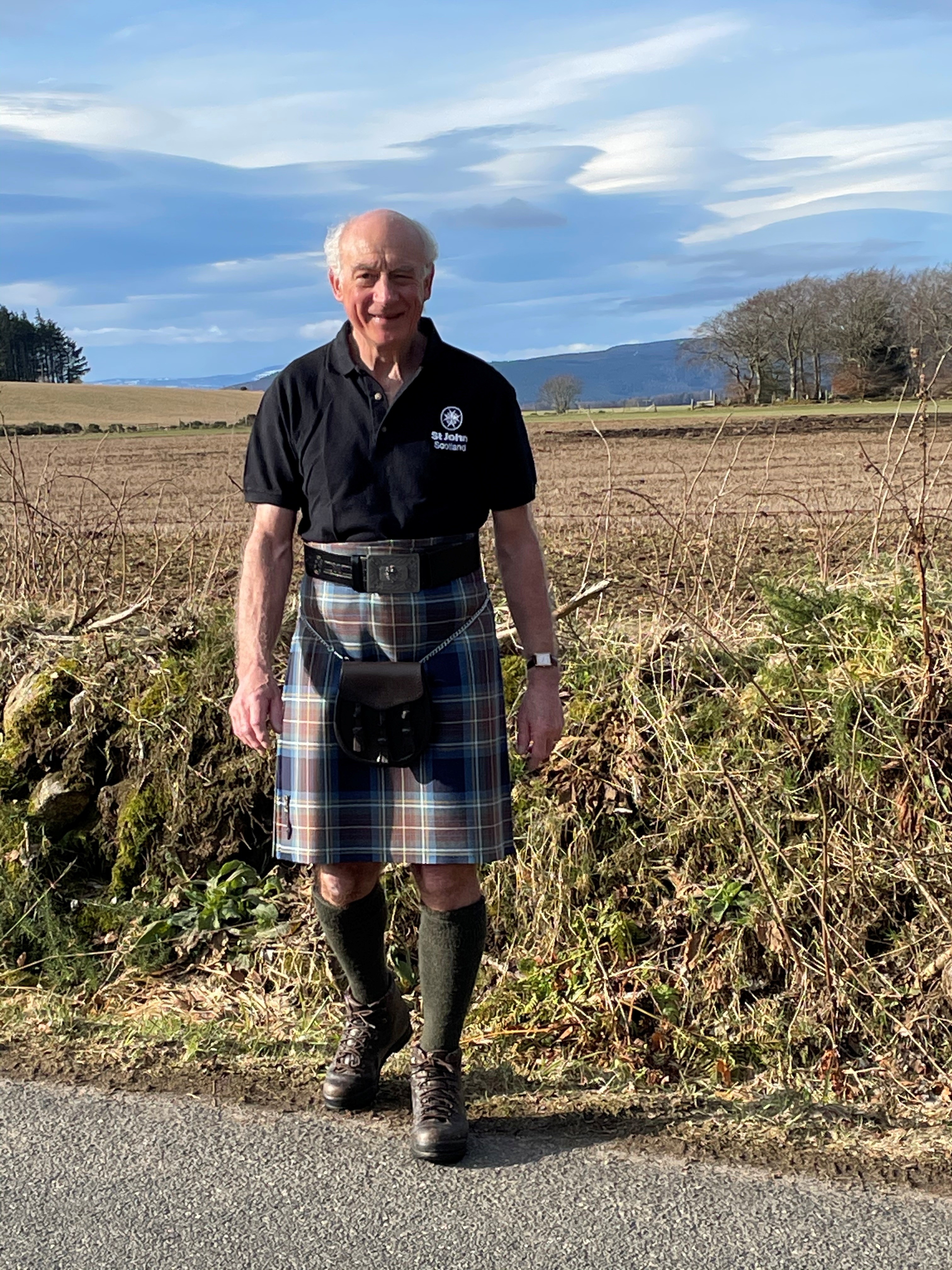 A man stands in front of a field with a St John Scotland polo shirt and a kilt on