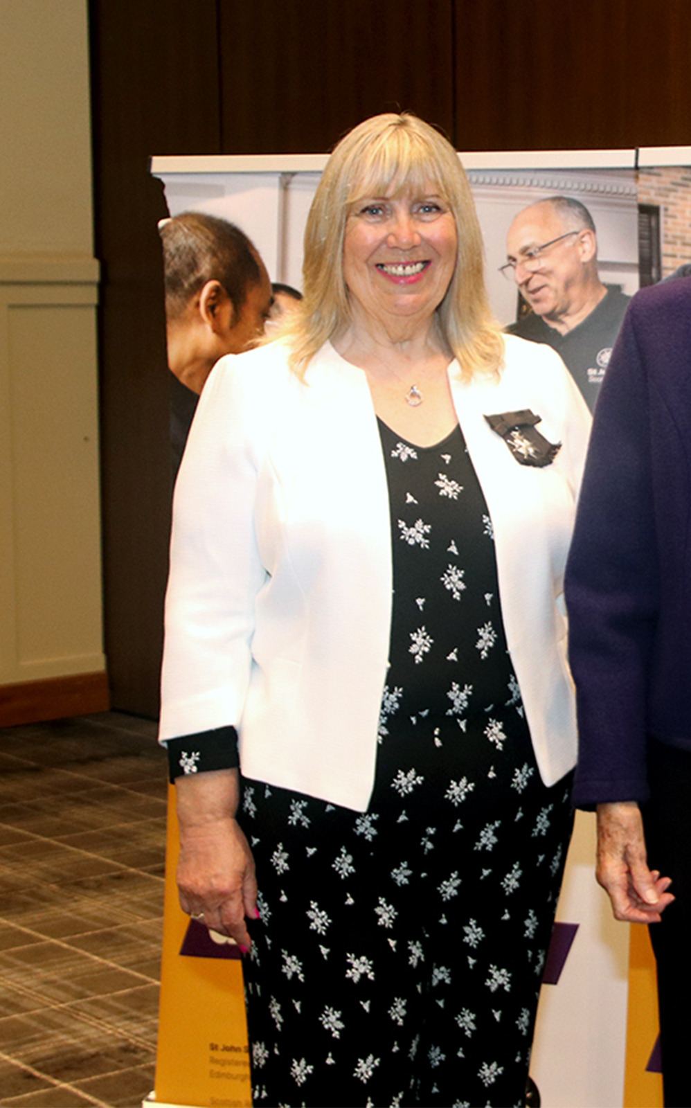 A blonde woman smiling in a black dress and white jacket