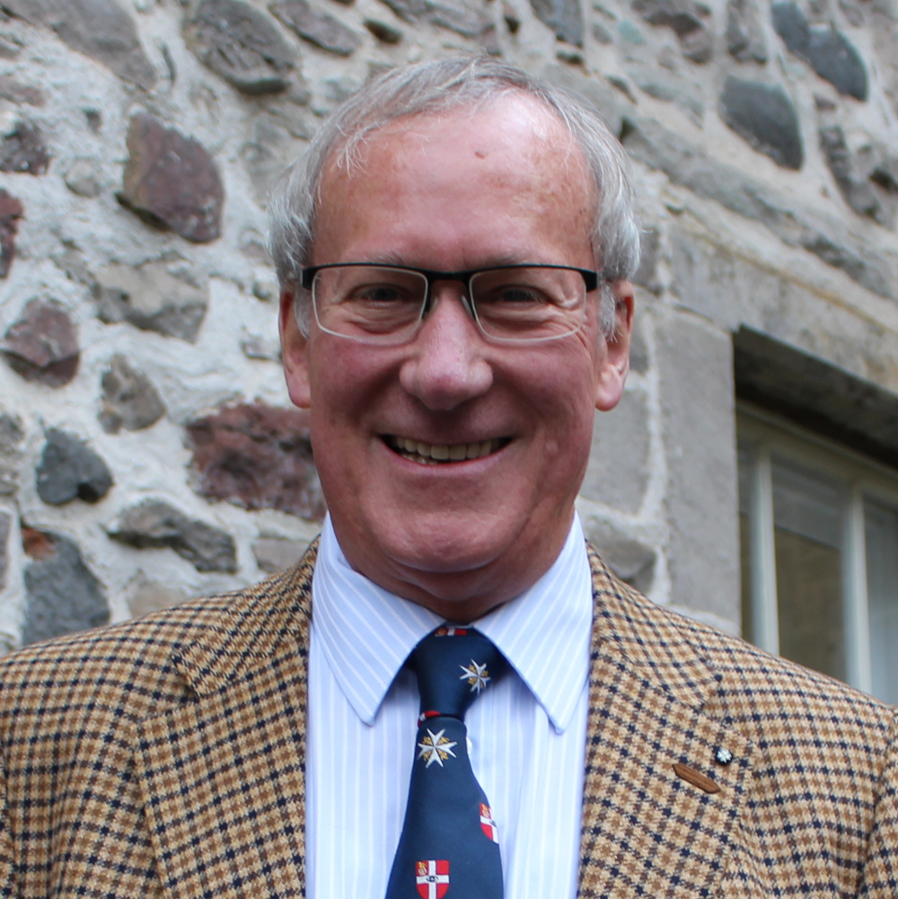 A man stands in front of a wall wearing glasses, a checked jacket and a St John tie